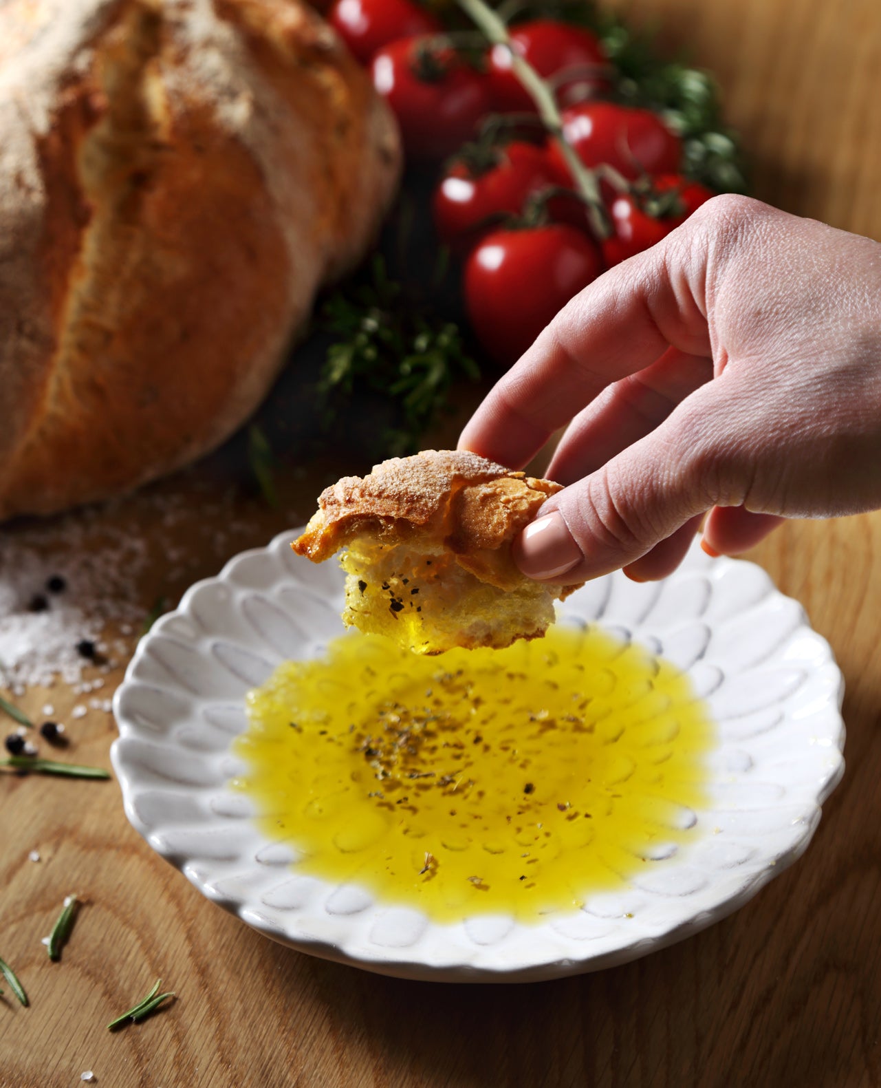 A kitchen table with a plate filled with olive oil and a hand dipping a slice of bread.
